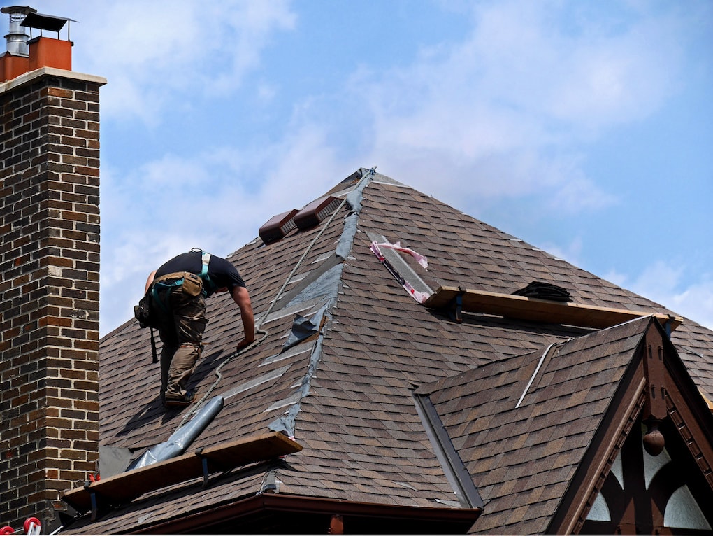 worker on roof replacing roof shingles against blue sky background